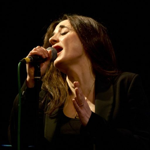 A photo of Laura Zakian, Jazz Singer. She is dressed in black and holding a microphone up to her mouth. She has her eyes closed and is singing with emotion.